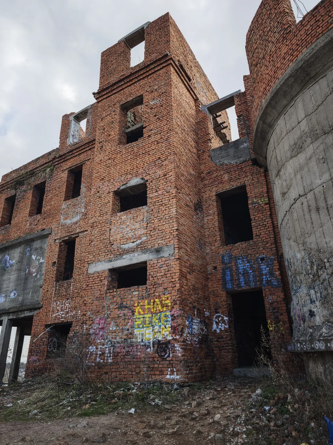 一座红砖建筑的废墟,墙上有多处窗户洞和涂鸦。建筑旁边有一个灰色的混凝土结构。天空阴沉,云层厚重。