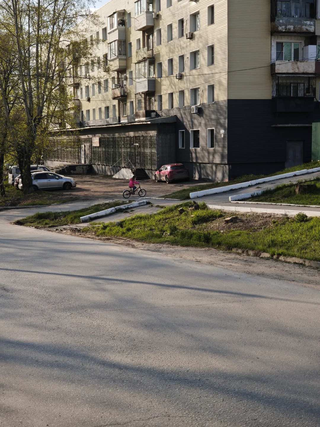 一条空旷的道路旁有一栋多层住宅楼,楼前停着几辆车。一个小孩正在骑自行车。树木在道路一侧生长。