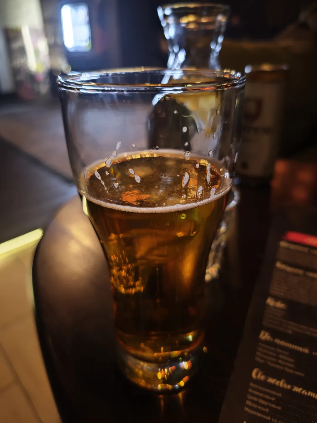 一杯金黄色啤酒放在桌面上,啤酒表面有泡沫。背景中可以看到菜单和其他餐具。玻璃杯反射出周围的光线。