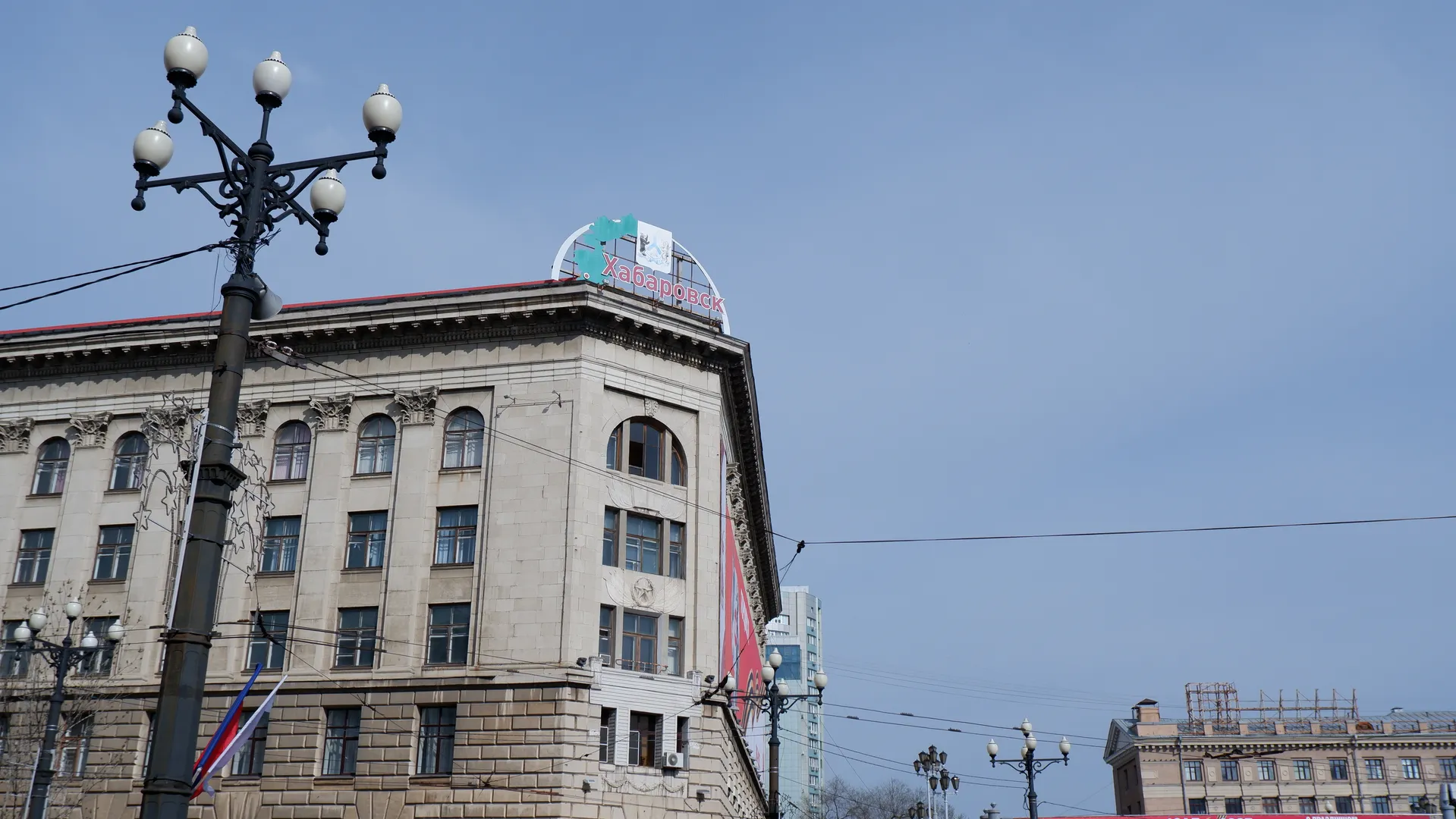 一座多层建筑的角落,顶部有“Хабаровск”字样。建筑前有路灯和电线。天空晴朗。