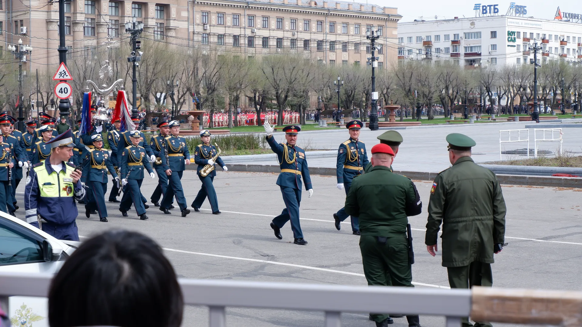 一支军乐队正在行进,成员们穿着深色制服并佩戴帽子。前方有两名身穿绿色军装的人员。背景是城市建筑和树木。