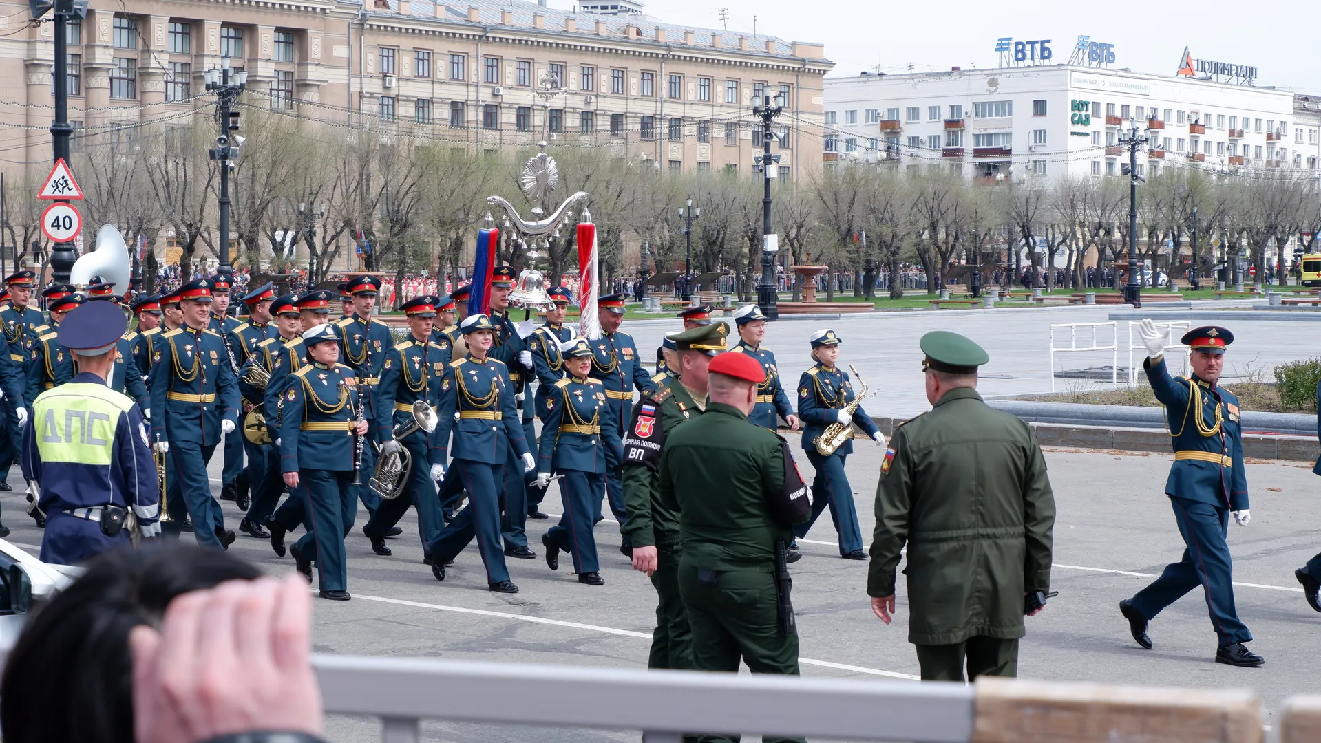 军乐队继续行进,成员们手持乐器,步伐一致。前方的绿色军装人员背对镜头。背景中的建筑物和树木清晰可见。