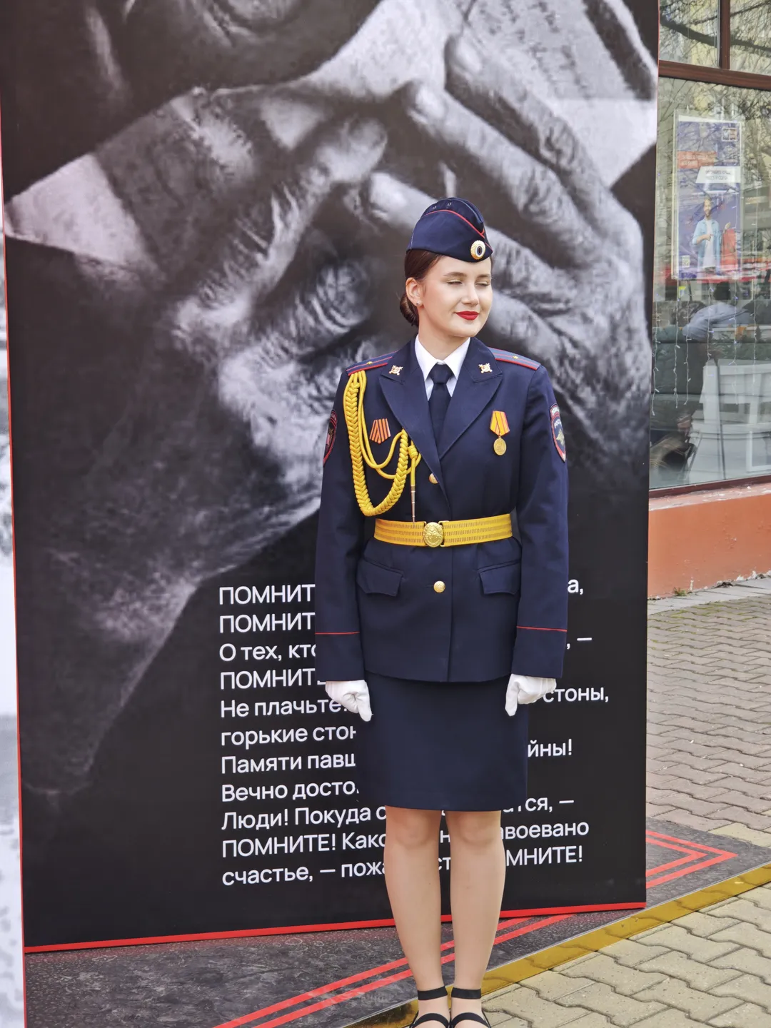 一位穿着深蓝色制服的女性站在海报前,制服上有黄色装饰带和徽章。海报上显示双手紧握的画面,并附有俄文文字。背景中可见玻璃窗和室内场景。