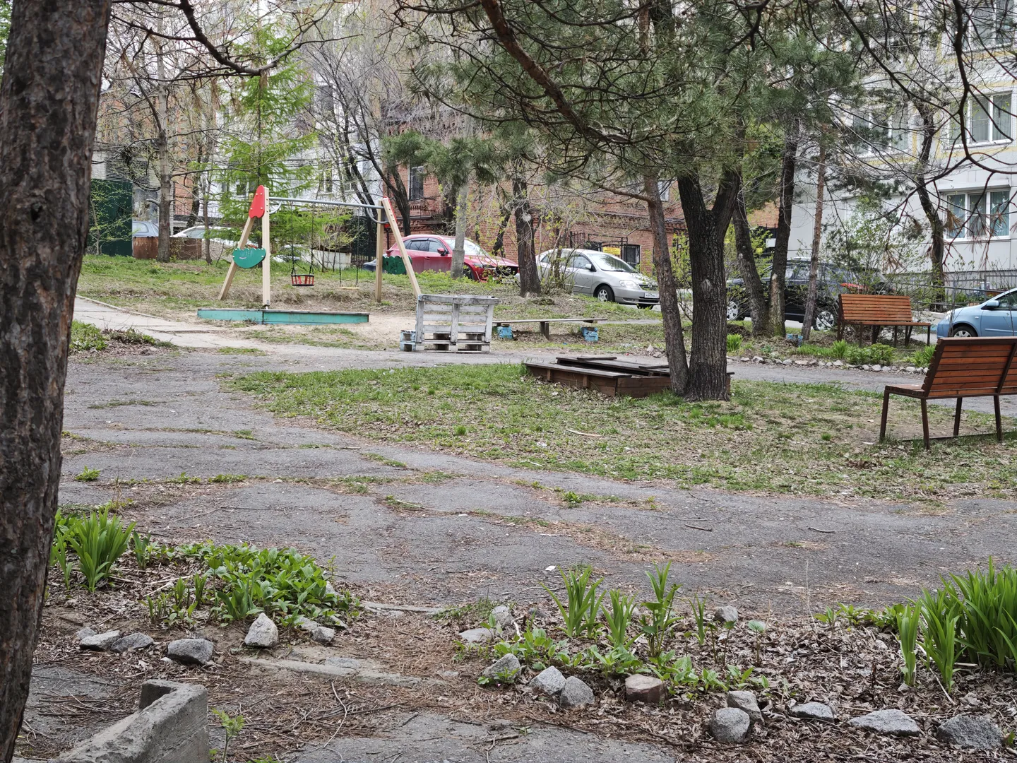 一片空旷的场地,地面部分覆盖着草和泥土,部分为硬化路面。场地上有几张木质长椅和儿童游乐设施。背景中停有多辆汽车。