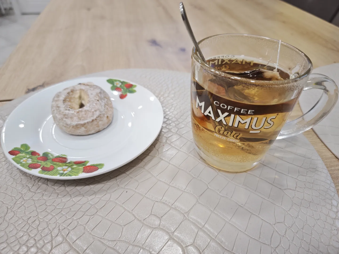 一张桌子上放着一个透明玻璃杯和一个白色盘子,玻璃杯里装有茶水,杯身上印有“COFFEE MAXIMUS Gold FREEZE-DRIED”字样。盘子里有一个撒有糖粉的甜甜圈。桌子表面有鳄鱼皮纹理。