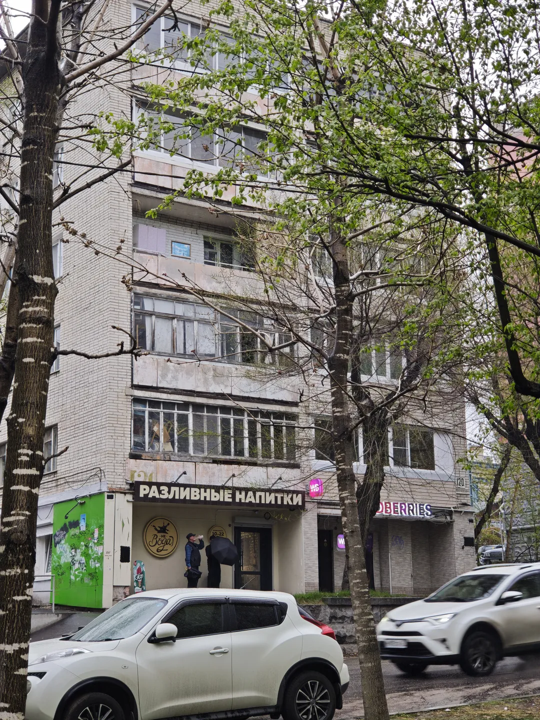 一栋多层建筑前停着几辆车,建筑底层有一家名为“РАЗЛИВНЫЕ НАПИТКИ”的店铺。树木的枝叶部分遮挡了建筑,街道上有行人和车辆经过。