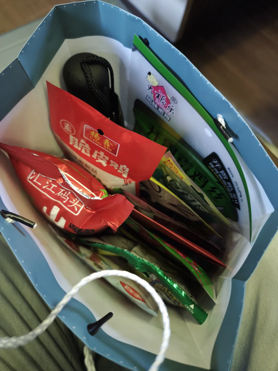 一个蓝色的纸袋里装有多种物品,包括红色和绿色包装的食品袋以及一个黑色的小物件。红色包装上印有“杨鑫脆皮鸡”的字样,绿色包装上有“开胃系列”的文字。袋子内部还放置了一些其他小包装食品。