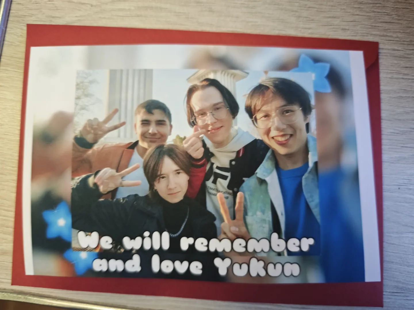 一张照片中有四个人,他们都比出胜利手势,照片下方写着“We will remember and love Yukun”。照片边缘装饰有红色边框和蓝色星星图案,背景中可见建筑物的部分结构。