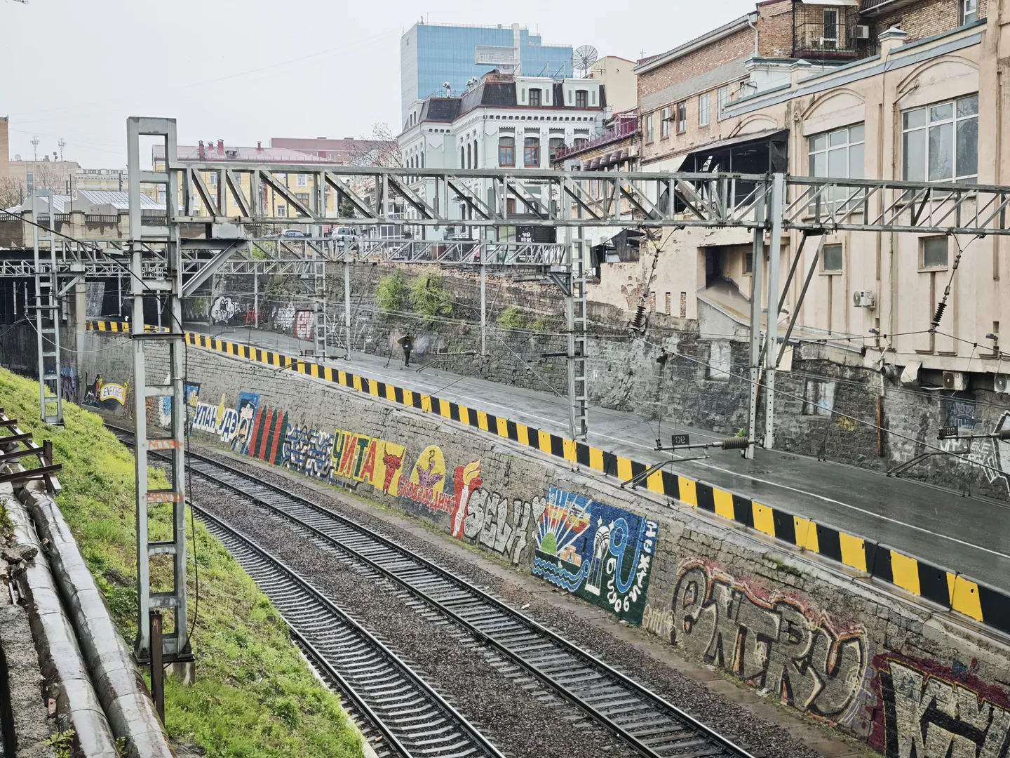 铁路轨道旁边有一条道路,道路上方有金属支架结构。轨道旁的墙上涂有彩色涂鸦,包括“ЧИТАТО”等字样。远处可以看到一些高层建筑和低矮的住宅楼。