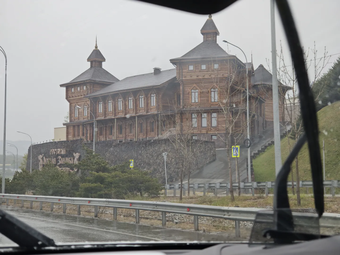 从车内视角看到一栋大型木质建筑,建筑有多层窗户和尖顶塔楼。建筑前方有护栏和几棵树,天空阴沉。车窗外的雨刷器部分遮挡了视线。