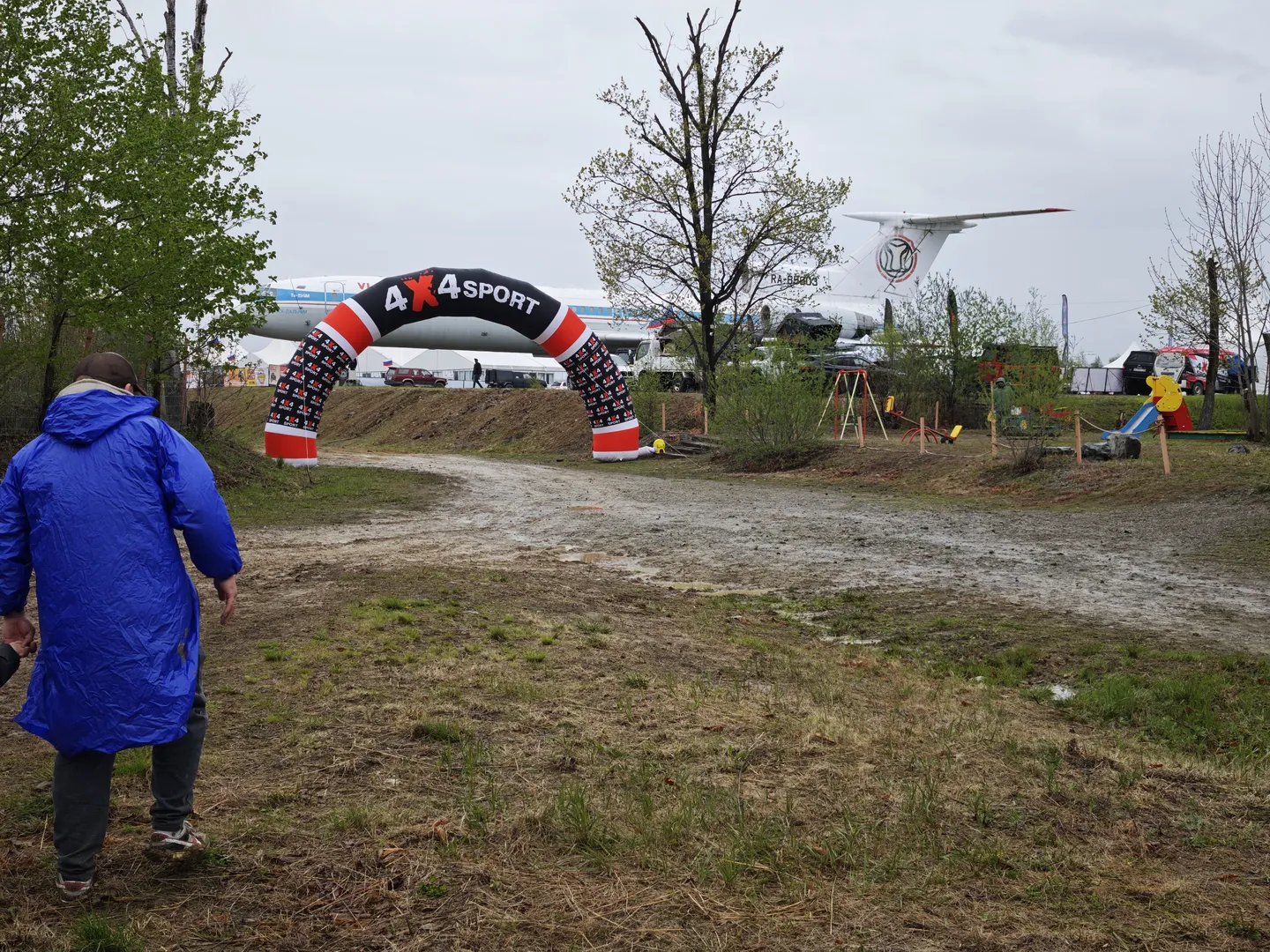 一个人穿着蓝色雨衣站在泥土地上,前方有一个标有“4x4SPORT”的拱门。背景中停着一架大型飞机,旁边有一些帐篷和游乐设施。地面湿滑,有明显的车辙痕迹。