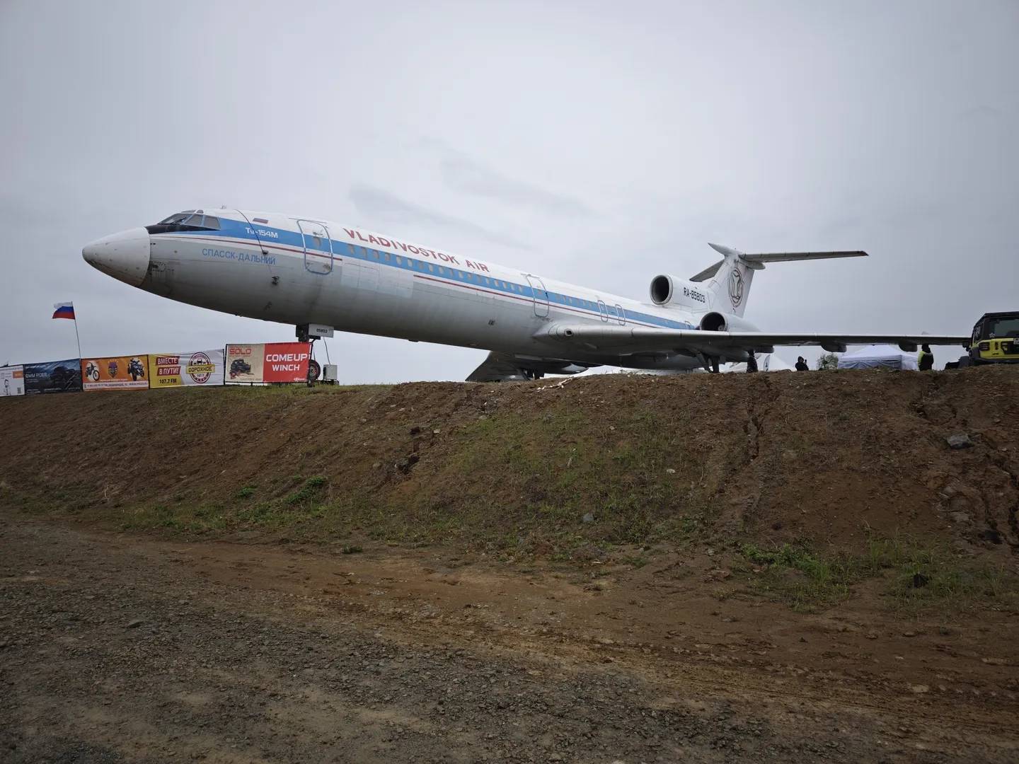 一架大型飞机停在土坡上,机身标有“VLADIVOSTOK AIR”字样。飞机前方有旗帜和广告牌,背景中有一些树木。地面为泥土,较为平坦。