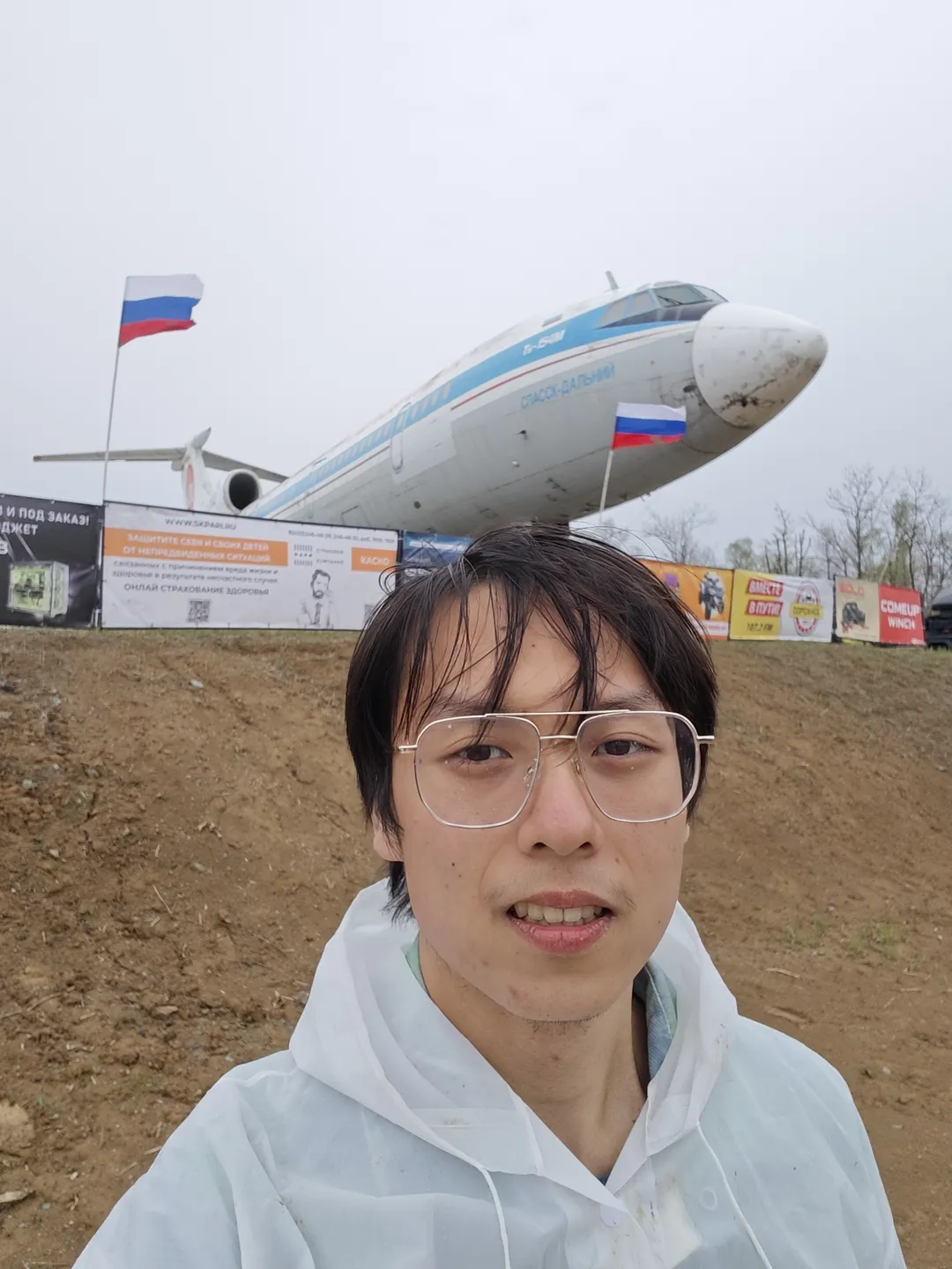 一个人穿着白色外套站在户外,背景中有一架大型飞机,机身上标有“VLADIVOSTOK AIR”字样。飞机前方有旗帜和广告牌,天空阴沉。地面为泥土,较为平坦。