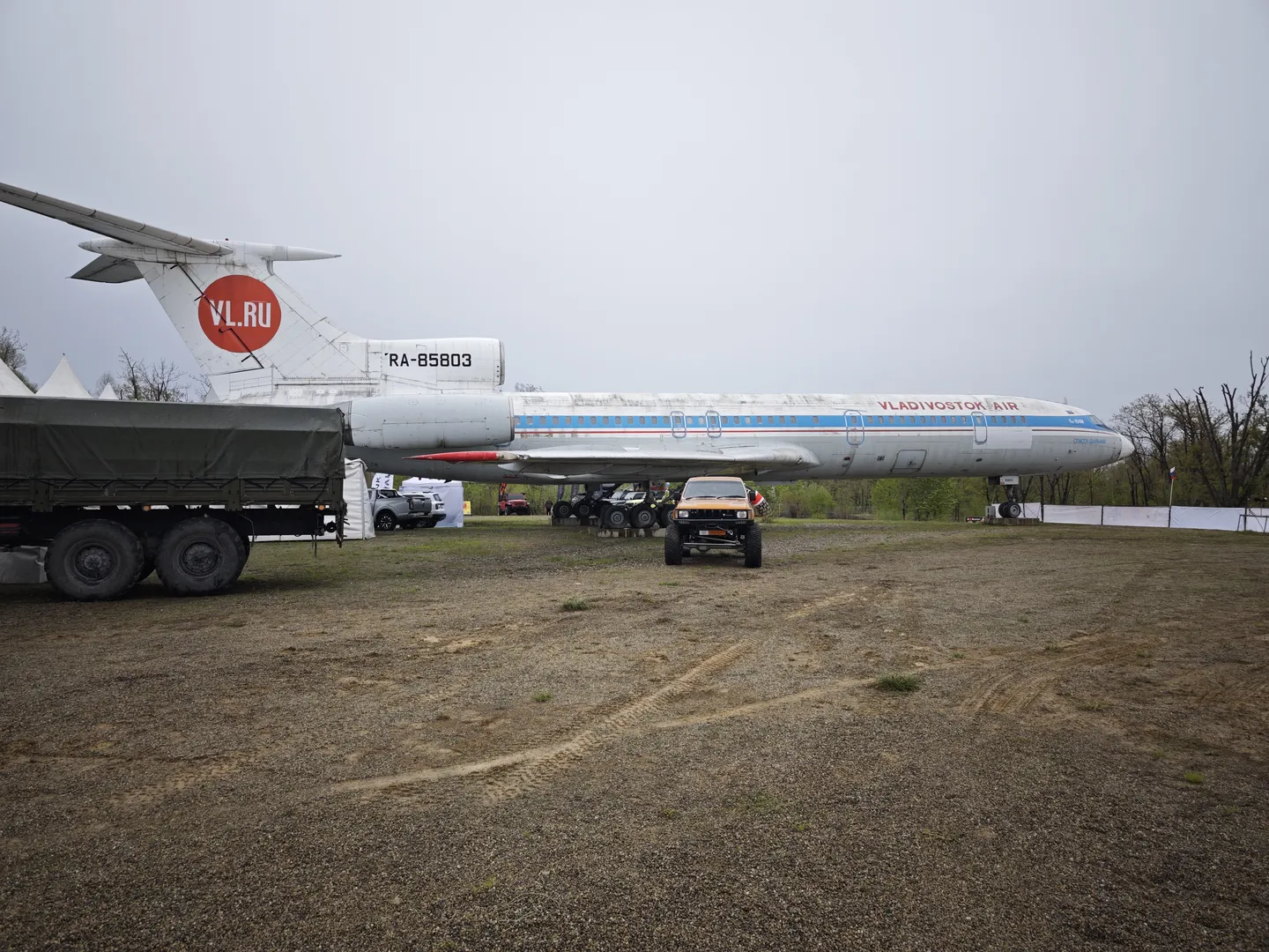 一架大型飞机停在空地上,机尾标有“VL.RU”字样。飞机前方停着一辆越野车和一辆卡车,背景中有树木和帐篷。地面为泥土,较为平坦。