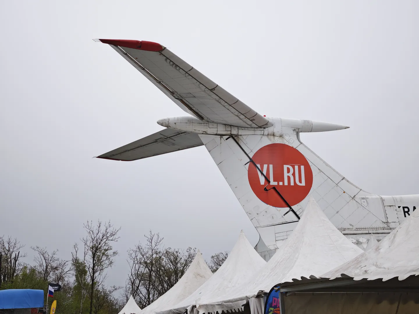 飞机尾部有一个红色圆形标志,上面写着“VL.RU”。飞机的尾翼和机身部分可见,背景是灰色天空。前景中有白色帐篷顶。