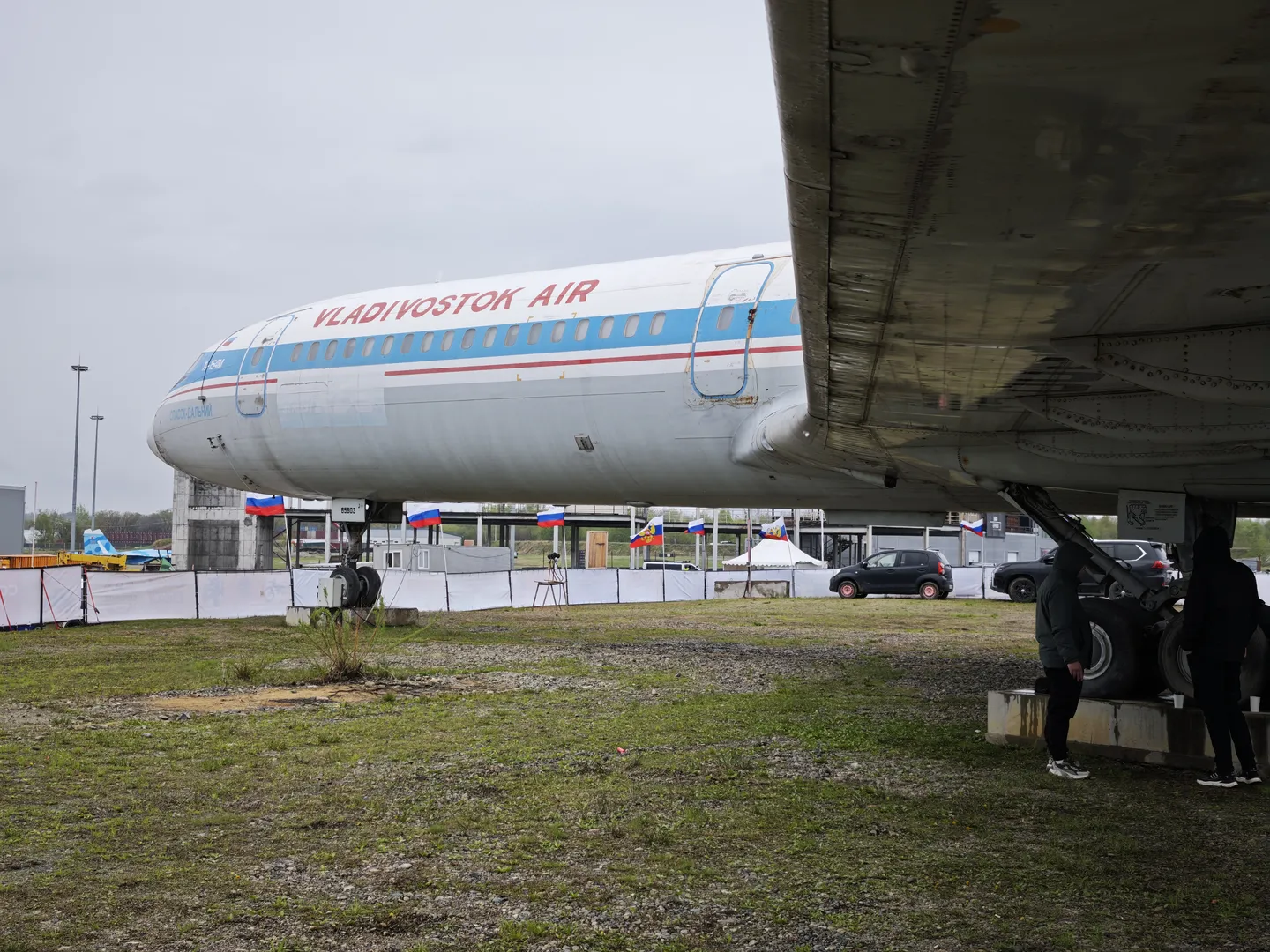 一架标有“VLADIVOSTOK AIR”的大型飞机停在草地上。飞机机身呈白色,带有蓝色和红色条纹。几个人站在飞机附近,背景有围栏和旗帜。