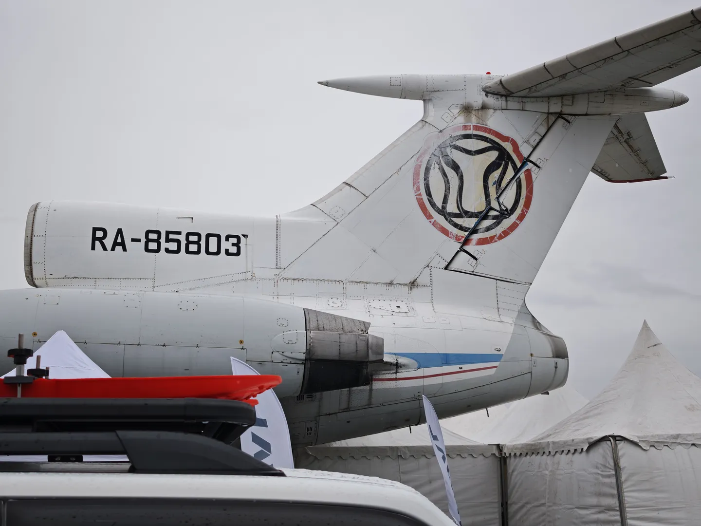 飞机尾部显示编号“RA-85803”,尾翼上有独特标志。飞机停在白色帐篷旁,背景为阴天。飞机引擎和尾翼细节清晰。