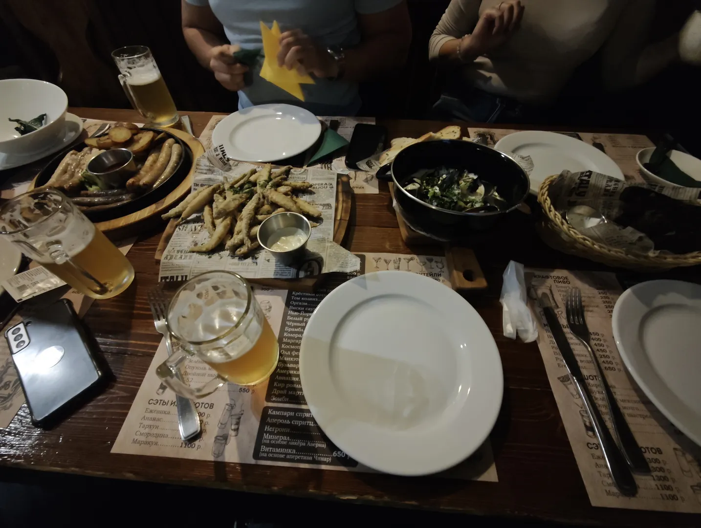 餐桌上有多种食物和饮料,包括啤酒、烤肠和沙拉。桌上还放有菜单和手机,几个人坐在桌旁。环境昏暗,灯光柔和。