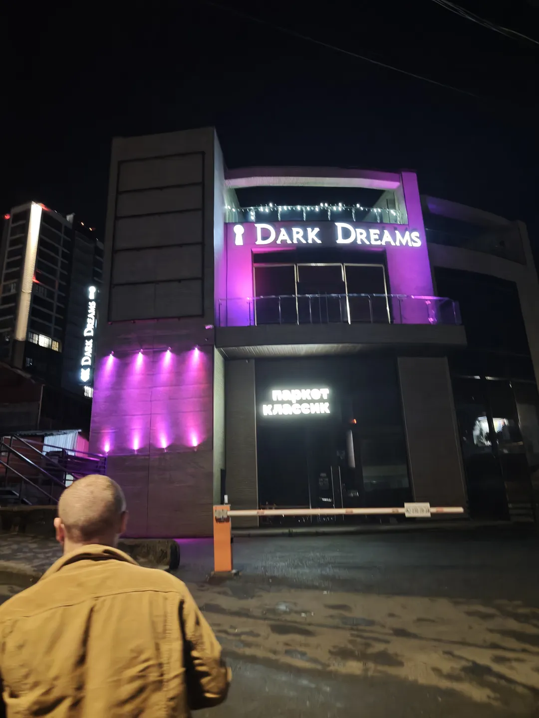 夜晚的建筑物外观,正面标有“Dark Dreams”字样,下方有“паркет классик”标识。建筑物外墙灯光呈紫色,门口有橙色栏杆。一名男子背对镜头站立。