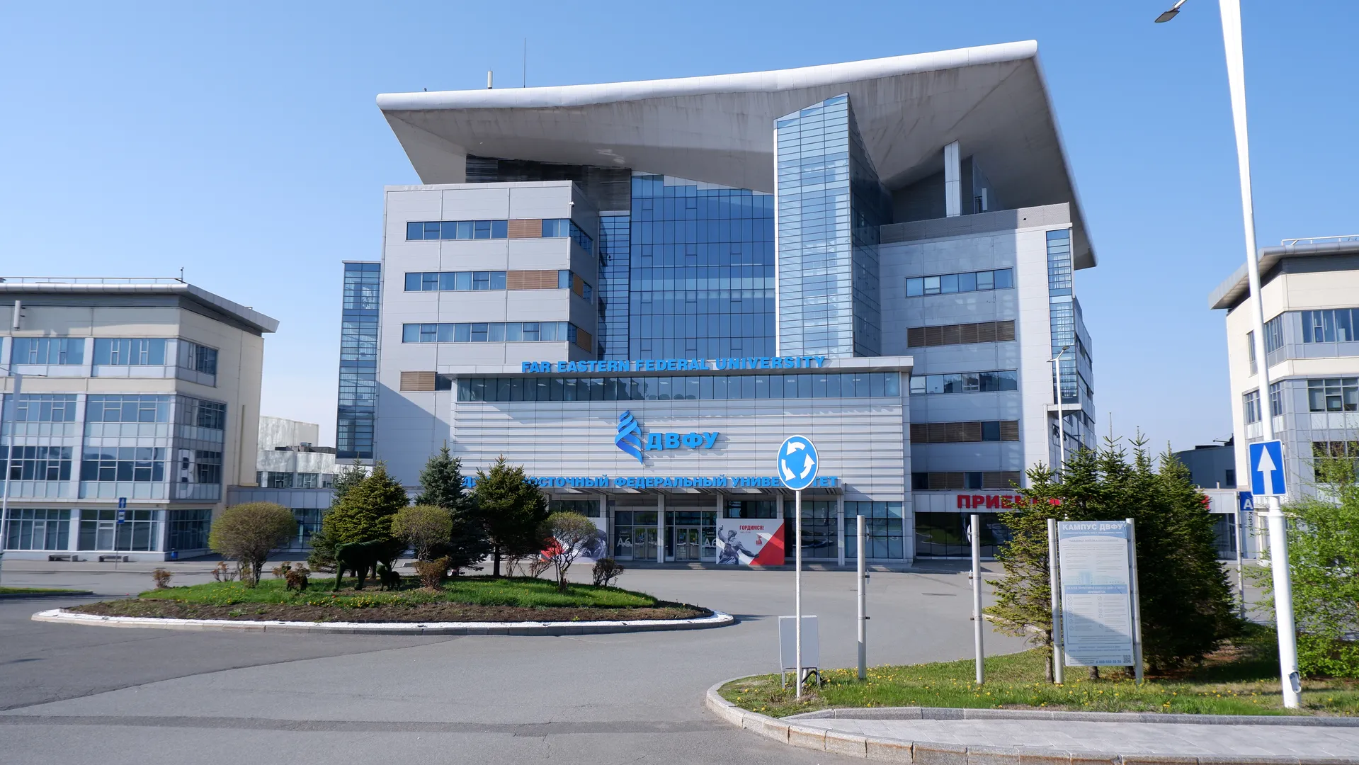 一栋高楼标有“FAR EASTERN FEDERAL UNIVERSITY”的字样,外墙由玻璃和金属构成,前面有绿化带和灌木丛。建筑物左侧有一栋较低的建筑,右侧则是一个指示牌。天空晴朗,背景中可见其他建筑物。
