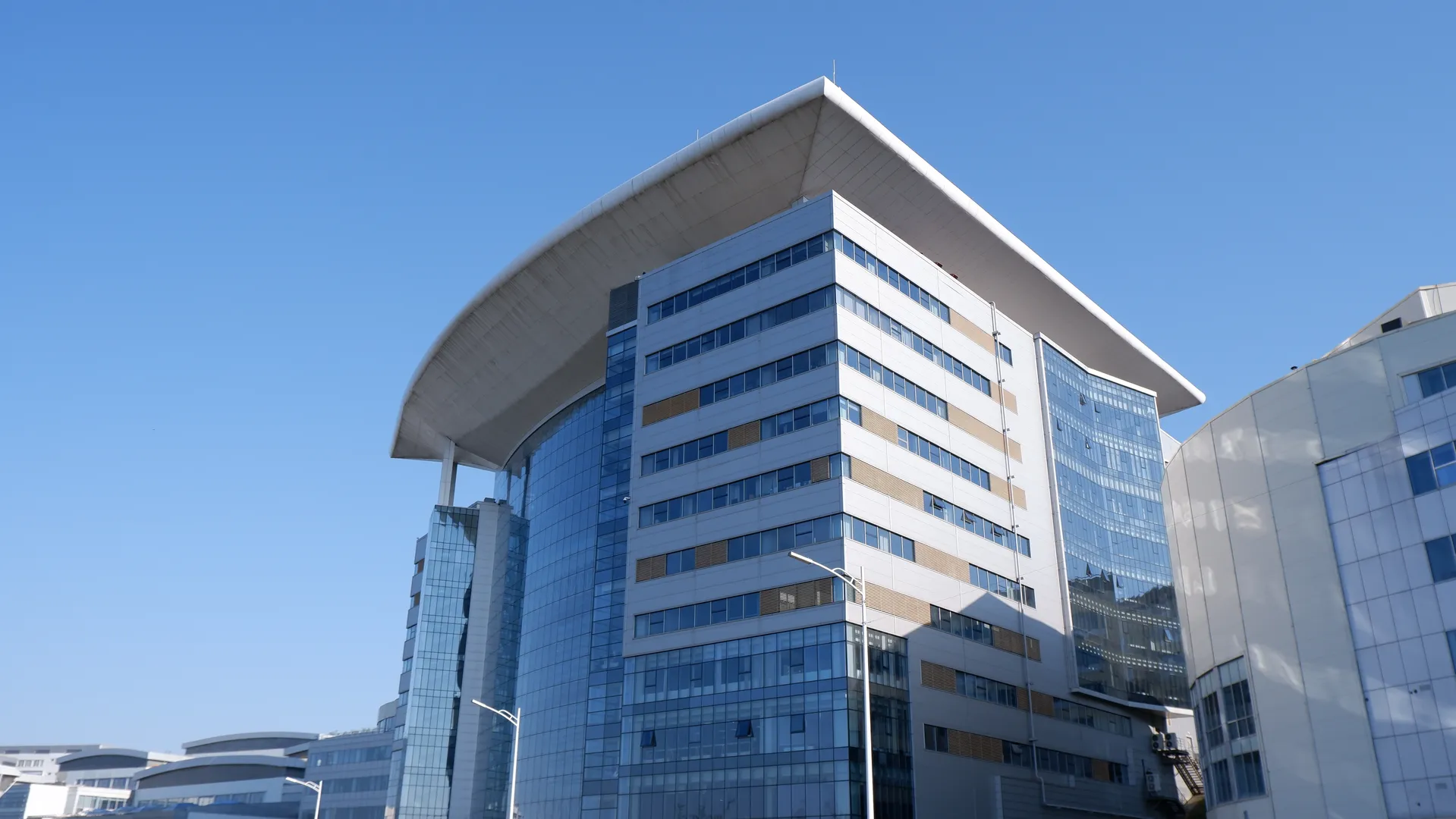 一栋现代化高层建筑,外墙采用大面积玻璃窗设计。建筑顶部有一个独特的弧形结构,反射着阳光。天空晴朗,背景中可以看到其他建筑物。