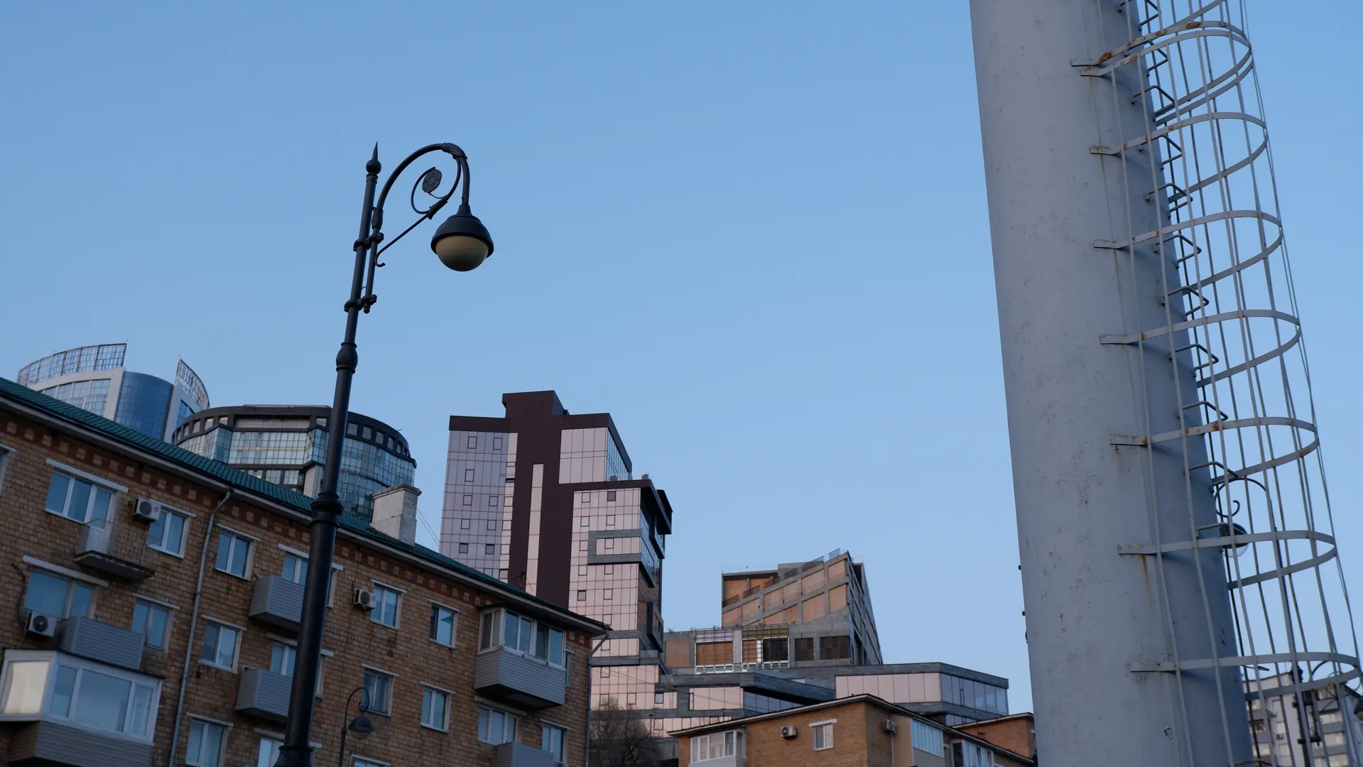 城市街道旁有一盏复古风格的路灯,背景中有多栋高层建筑。建筑物的设计现代,外墙有玻璃幕墙。天空晴朗,呈现出淡蓝色。