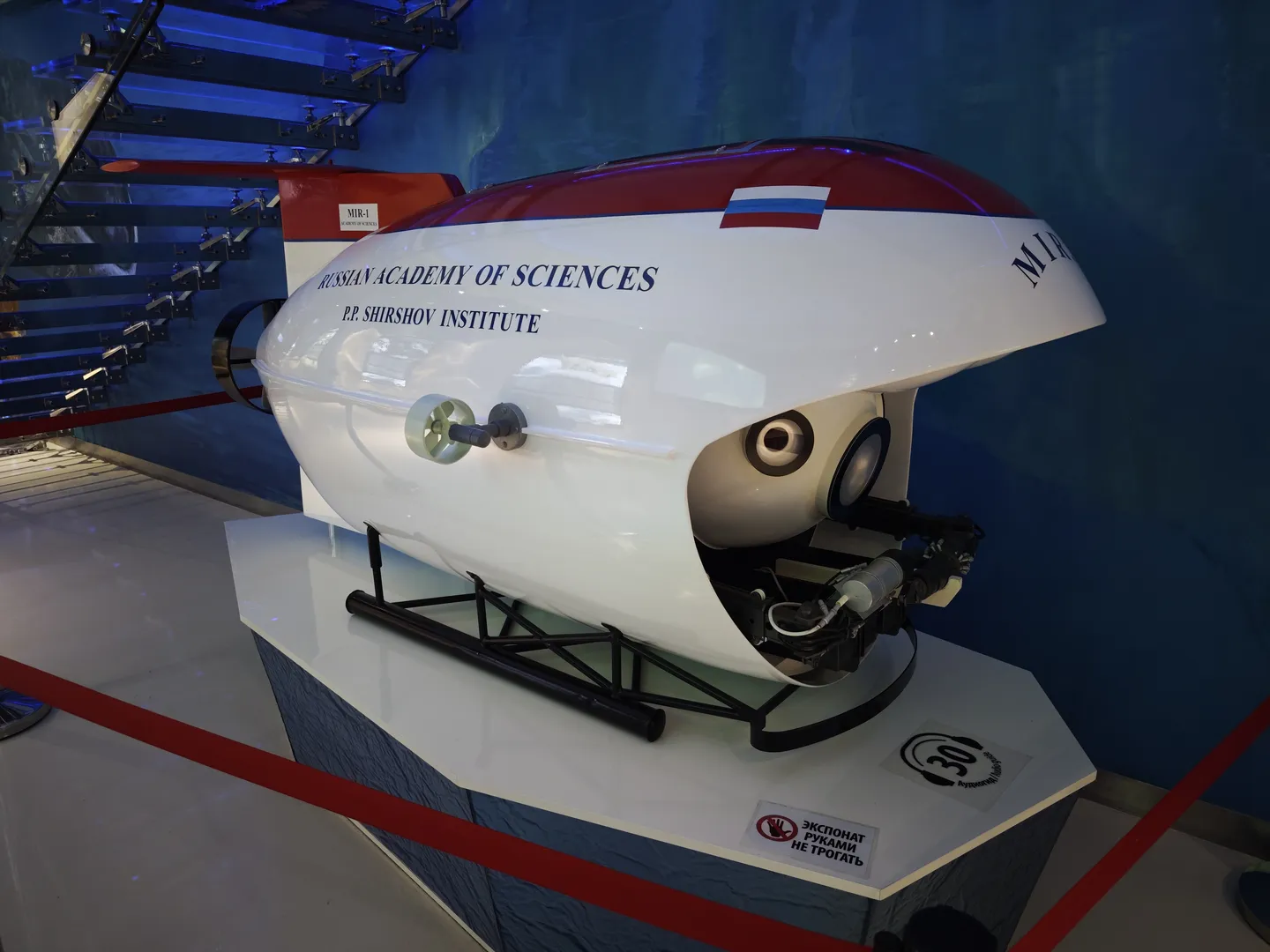 一台标有“RUSSIAN ACADEMY OF SCIENCES P.P. SHIRSHOV INSTITUTE”的潜水器模型放置在展台上。模型呈流线型设计,主体为白色,带有红色和蓝色条纹。旁边有禁止触摸的标志。