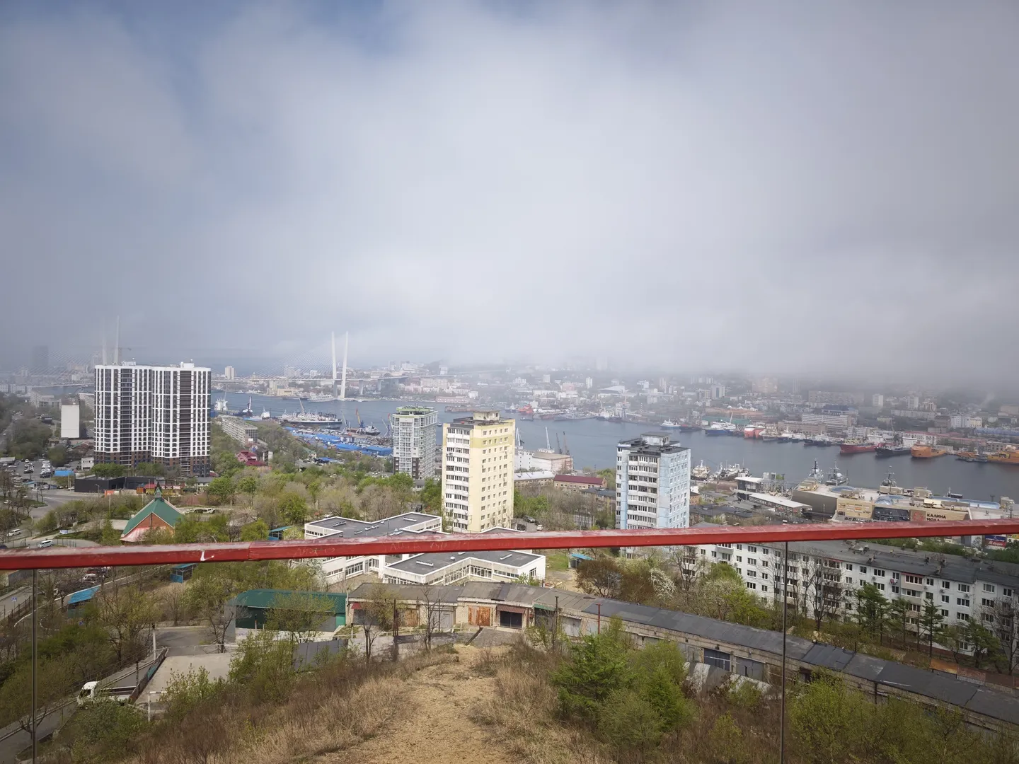 城市全景从高处俯瞰,前景是低矮建筑和绿植,中景是高层建筑和河流,河上有桥梁和船只。天空阴沉,云雾缭绕,遮挡了部分视线。前景有红色护栏。