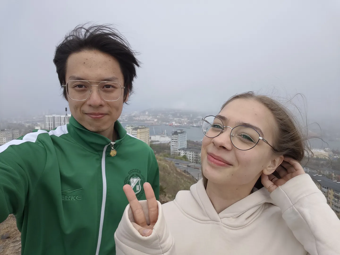 两个人站在高处自拍,背景是城市景观和海港。左边的人穿着绿色夹克,右边的人穿着白色连帽衫,两人面带微笑。