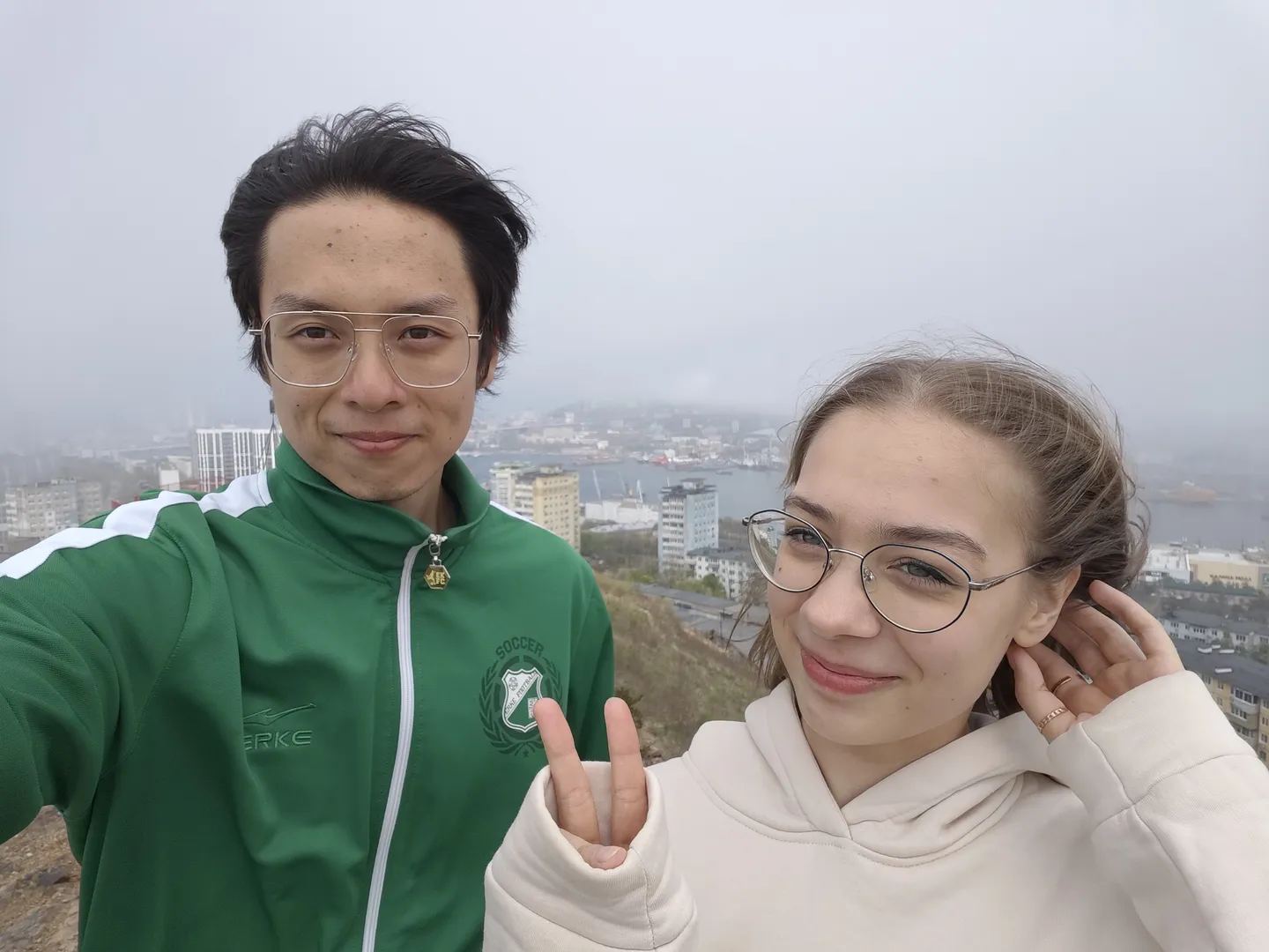 两个人站在高处自拍,背景是城市景观和海港。左边的人穿着绿色夹克,右边的人穿着白色连帽衫,两人面带微笑。