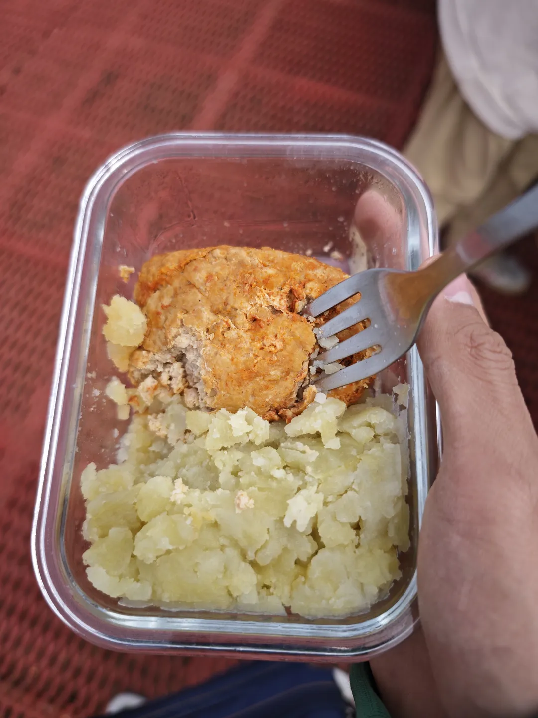 一只手拿着叉子,叉子插在一个透明容器中的食物上,容器里有土豆泥和一块炸肉饼。容器放在红色桌布上,叉子正准备叉起食物。
