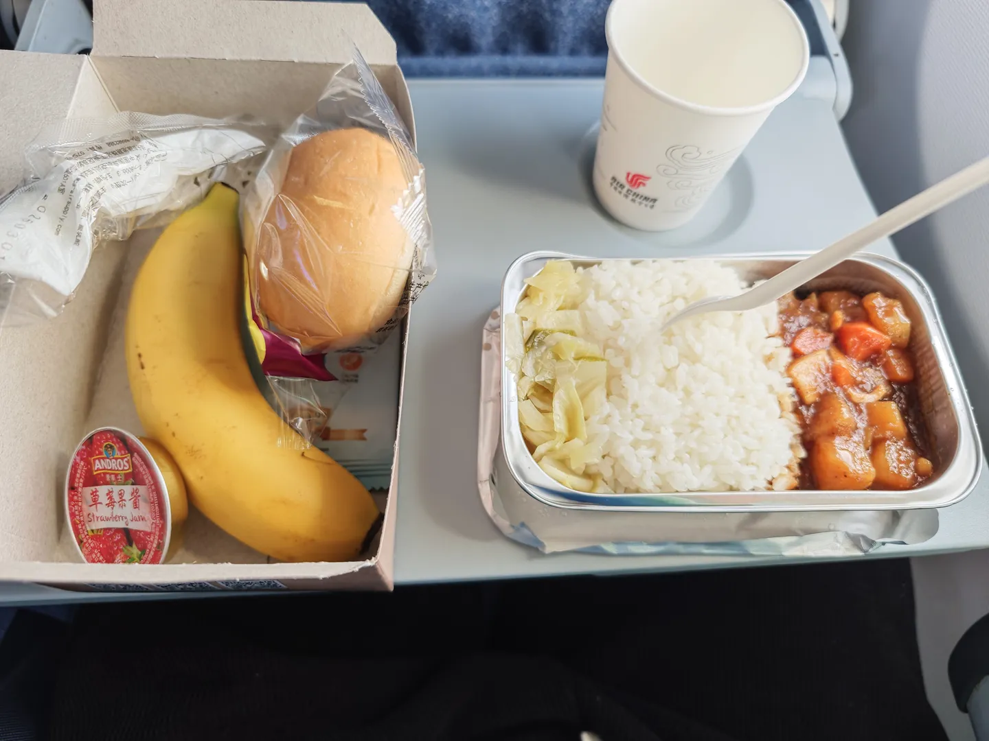 飞机餐盘中包含一份米饭、炖菜和泡菜,旁边是一个香蕉、一个面包和一小罐草莓果酱。还有一个纸杯,上面印有“Air China”的标志。