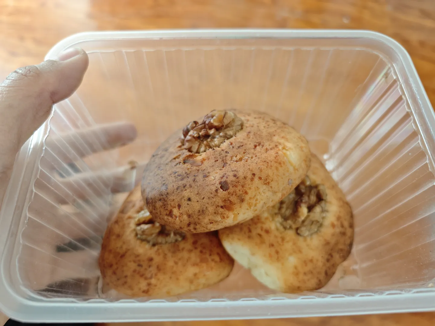 透明塑料盒内装有三个圆形糕点,表面撒有坚果碎屑。糕点颜色金黄,形状规整。一只手正拿着塑料盒。