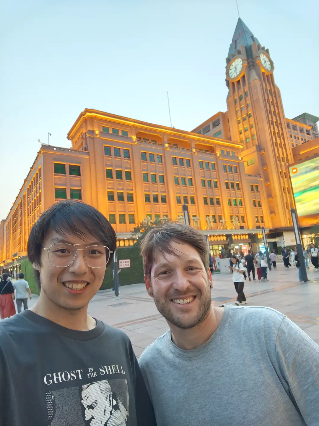 两个人站在一座大型建筑前,建筑上有明亮的灯光和一个高大的钟塔。建筑的外墙呈黄色,窗户排列整齐。背景中有一些行人和绿色植物。