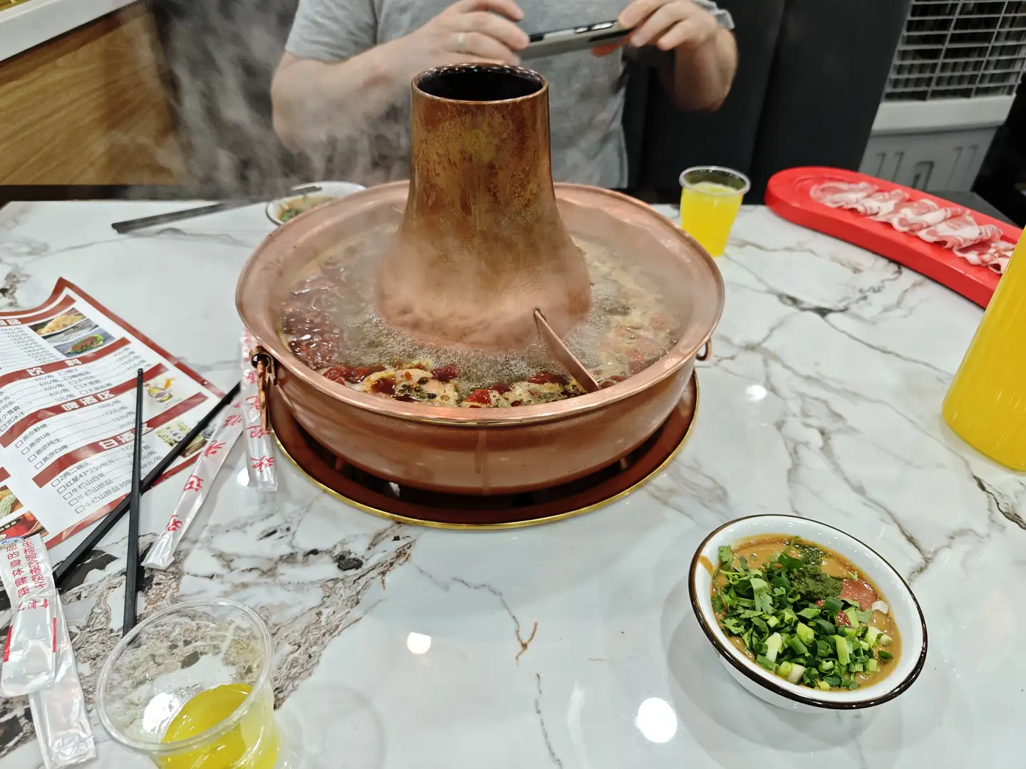 桌子上有一个铜制火锅,锅内有热气升起,旁边放着一碗蘸料和一些餐具。火锅分为两个部分,一侧有红色辣椒。背景中有人正在用手机拍照。