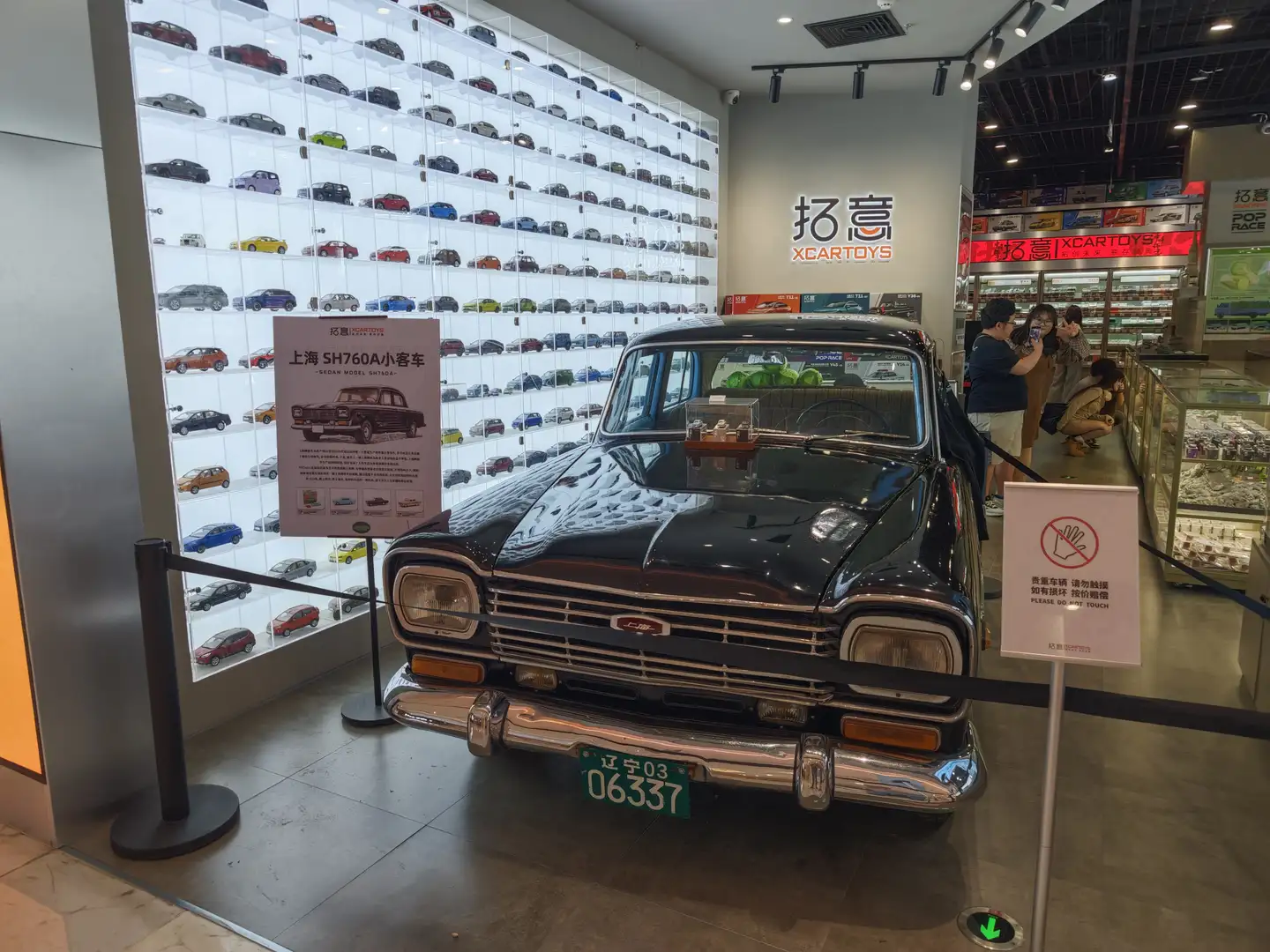 展厅内展示了一辆黑色复古汽车,车牌号为辽宁03 06337,旁边有一个标牌介绍汽车信息。墙上陈列着许多小型汽车模型,形成一面汽车墙。展厅内还有几位参观者在观看展品。