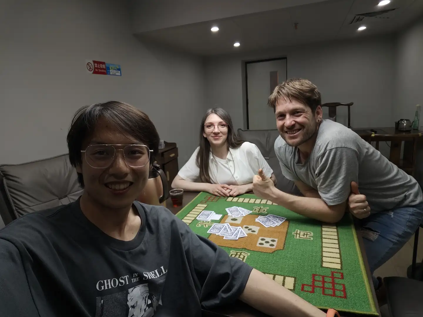 三个人坐在桌子旁,桌上铺着绿色麻将桌布,上面摆放着麻将牌。左边的人穿着印有“GHOST IN THE SHELL”字样的黑色T恤,右边的人穿着灰色T恤,中间的人穿着白色上衣。背景是一个室内房间,墙上贴有禁止吸烟标志。