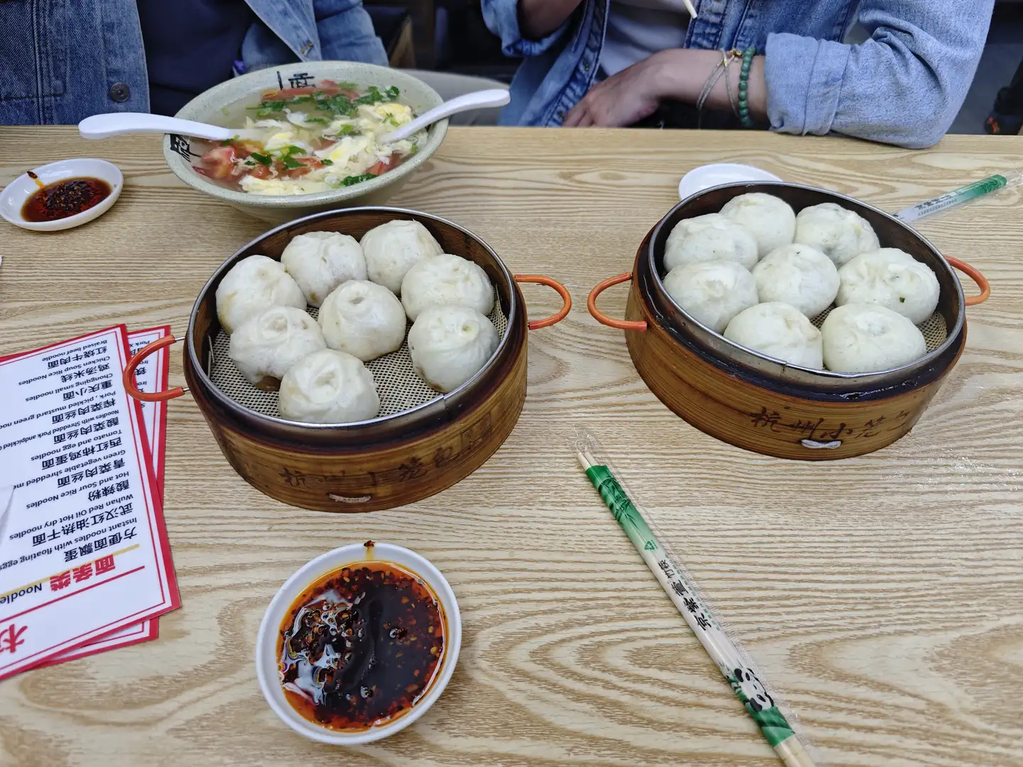 桌上摆放着两笼包子、一碗汤和一些调料。旁边有一双筷子和菜单,菜单上列有各种面食选项。桌子是木质的,表面有纹理。
