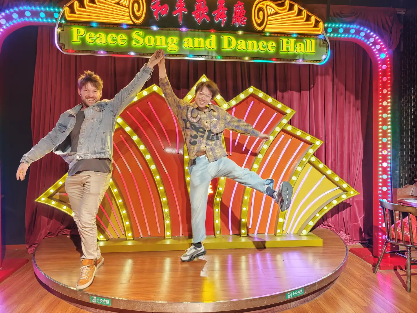 两个人站在舞台上,舞台背景标有“Peace Song and Dance Hall”的霓虹灯牌。左边的人穿着浅色衣服,右边的人单腿抬起,穿着蓝色裤子。舞台灯光闪烁。