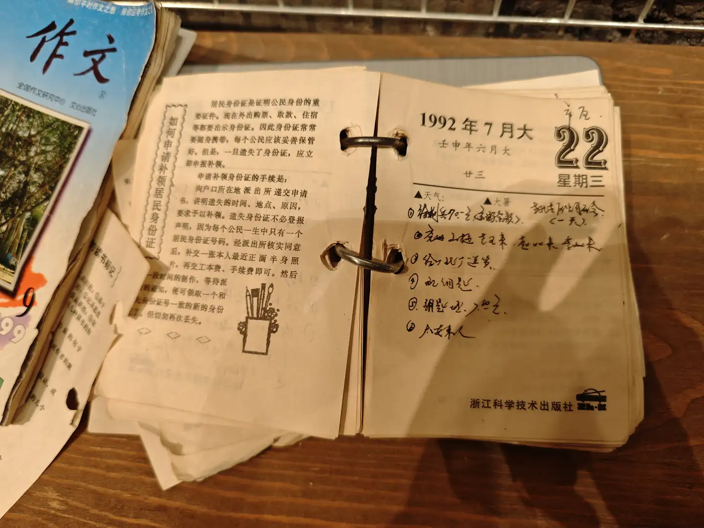 一本打开的日历显示1992年7月22日,旁边有一本作文书。日历上记录了当天的天气和几项活动。页面设计简洁,字体清晰。
