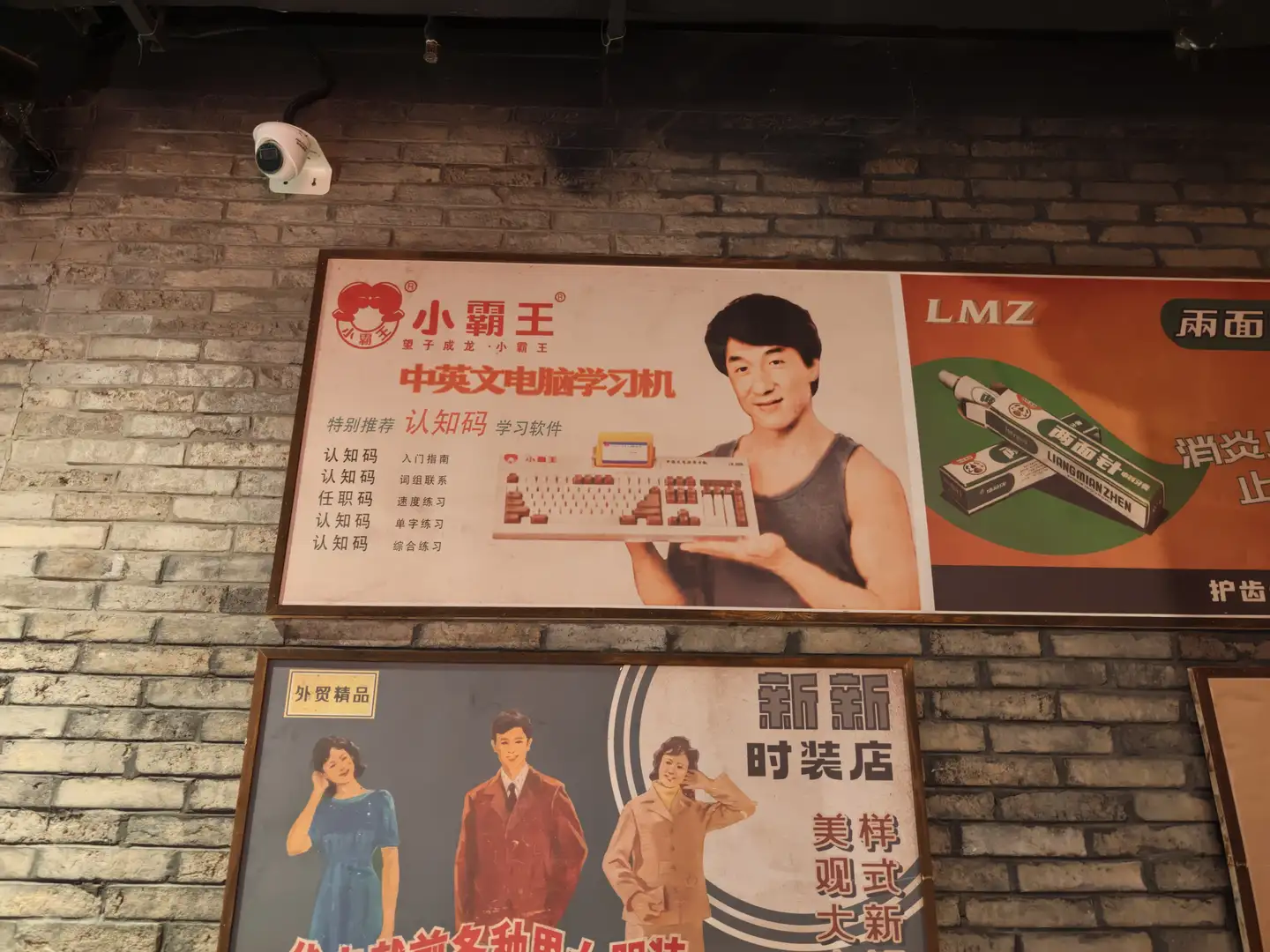 墙上挂着两幅广告海报,上方海报展示“小霸王 中英文电脑学习机”,下方海报宣传时装店。海报色彩鲜艳,人物形象生动。背景为砖墙,上方有监控摄像头。
