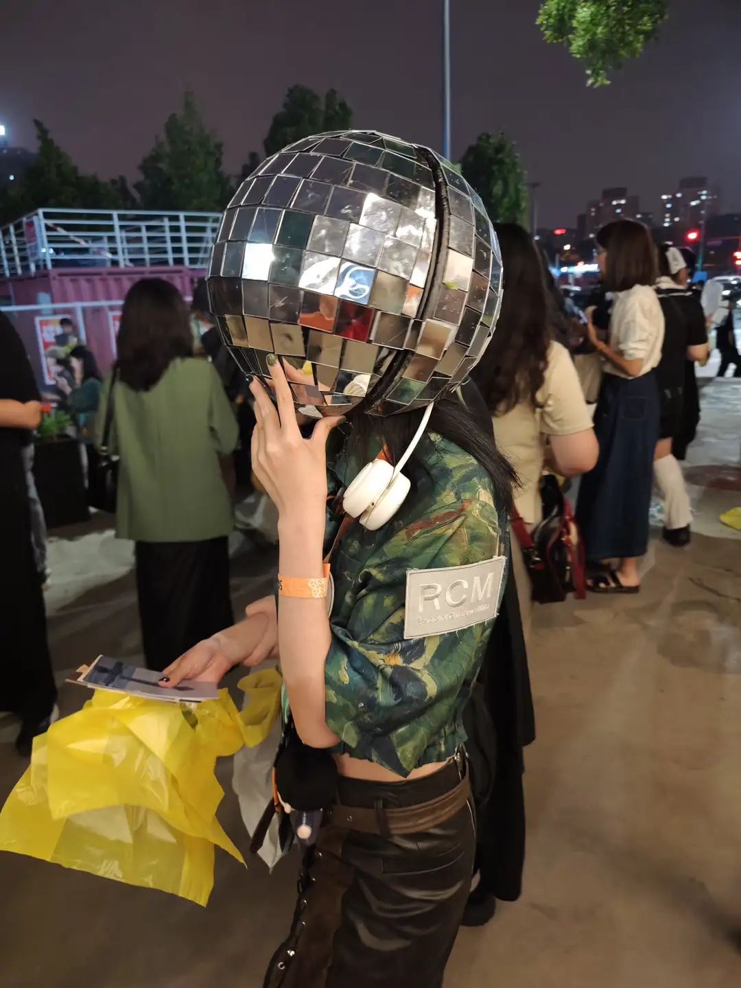 一个人戴着由镜子组成的球形头盔,穿着迷彩上衣和黑色裤子,手里拿着黄色塑料袋。背景中有多个人在行走,场景似乎是在夜晚的户外。