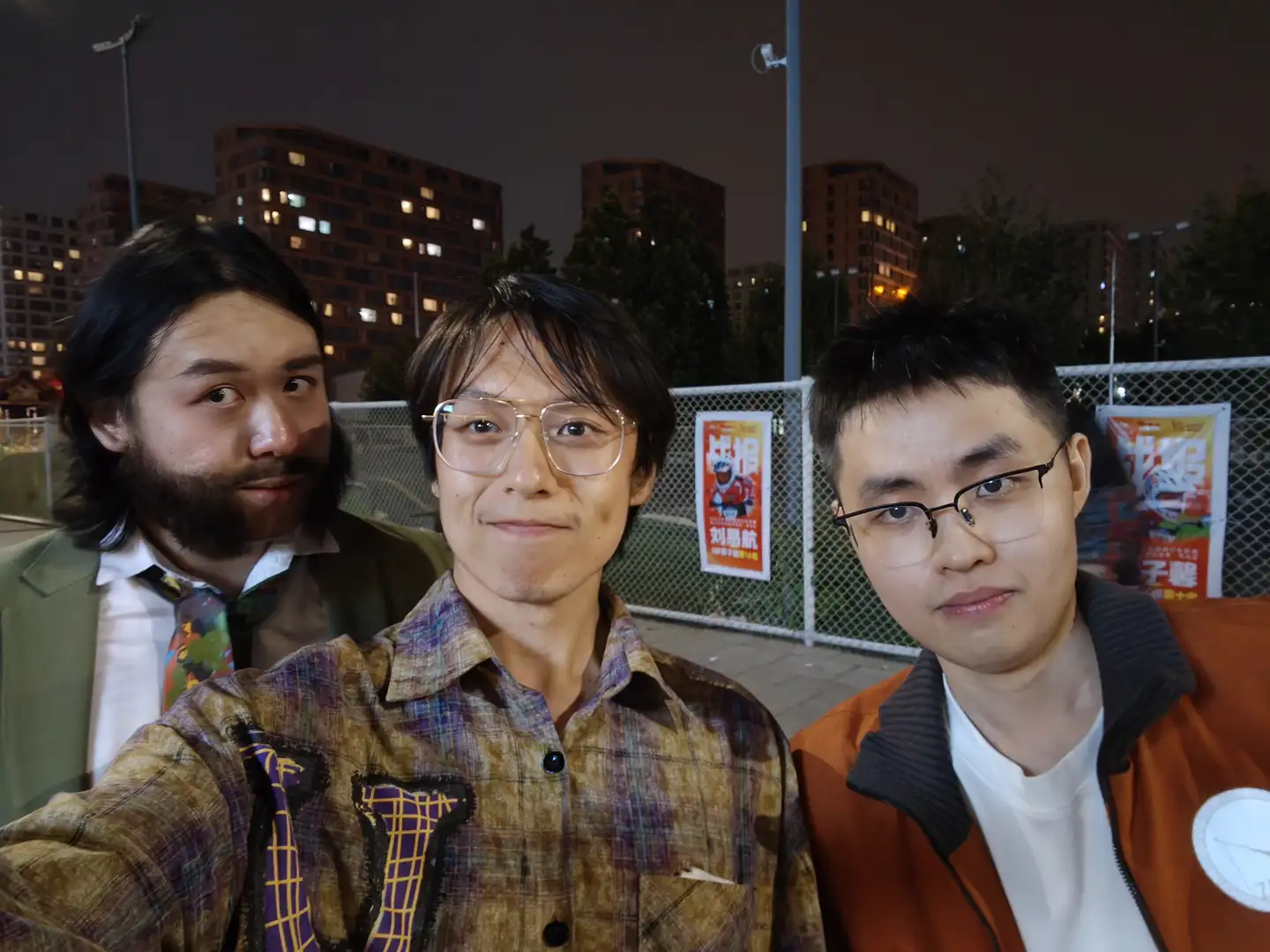 三个人站在一起自拍,左边的人穿着格子衬衫,中间的人穿着相同风格的衬衫,右边的人穿着橙色外套。背景是夜晚的城市景观和铁丝网围栏,围栏上有张贴的海报。
