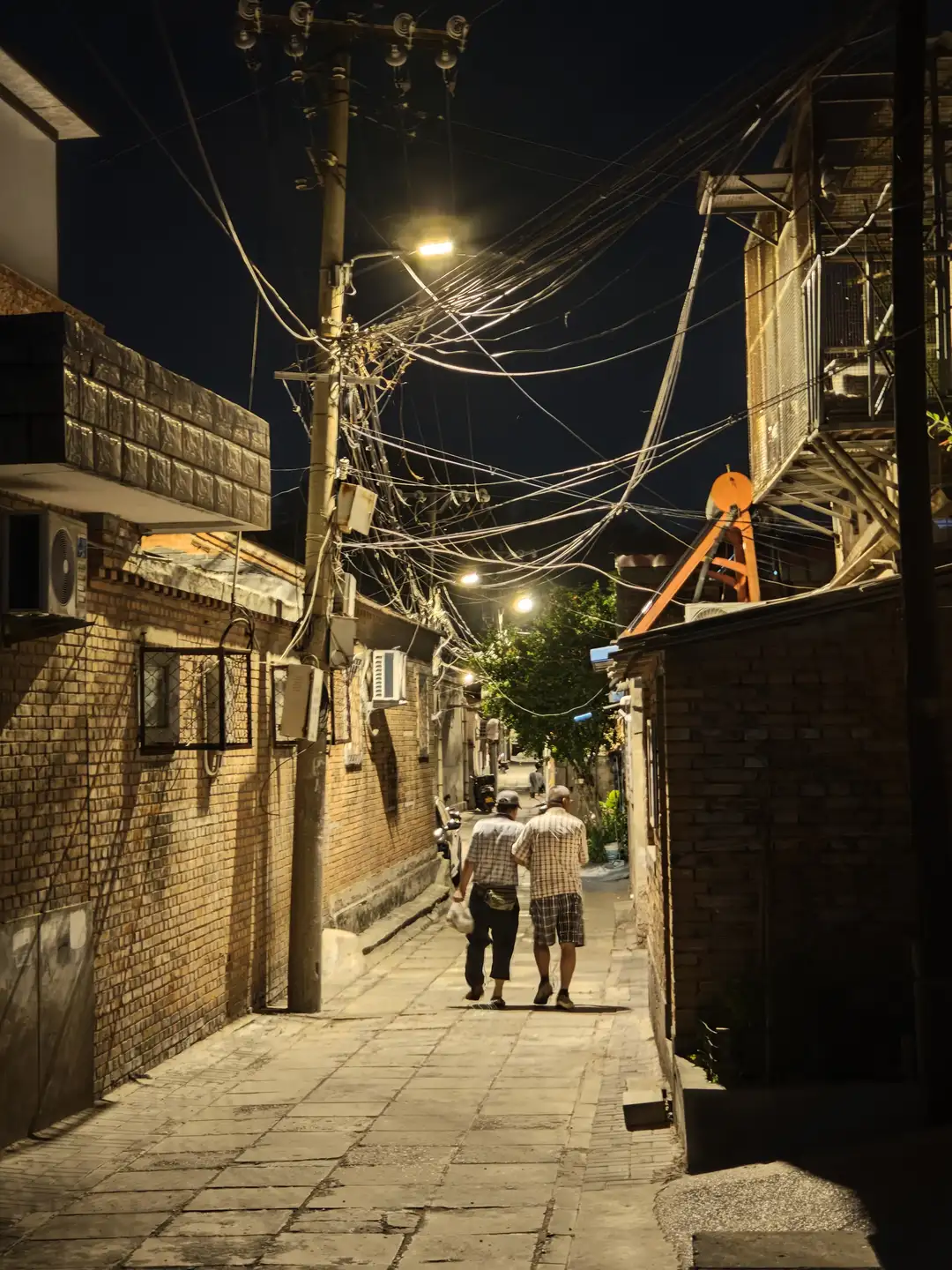 夜晚的巷子里,两个人并肩行走,周围是砖墙和电线杆。路灯照亮了他们的前方,电线在空中交错。远处有树木和更多的建筑物。