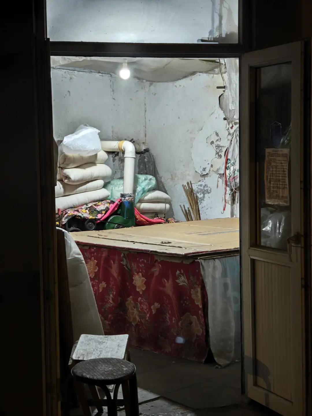 房间内部陈设简单,墙上剥落的油漆显露出岁月痕迹。门口堆放着被褥和杂物,中央有一张覆盖红色花纹布料的桌子。墙上贴着一张纸,内容为注意事项或公告。