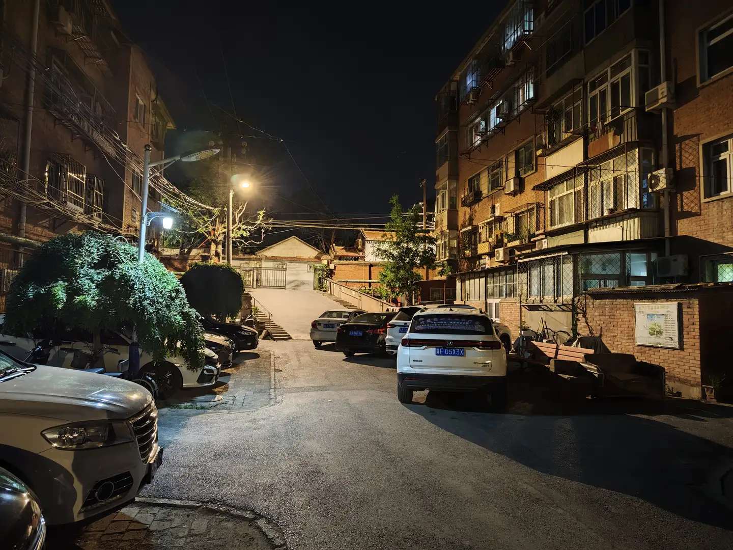 夜晚的住宅区,街道两侧停满了车辆,路灯照亮了道路。建筑物多层,阳台上装有防护网和空调外机。街道尽头有一段台阶和围栏。