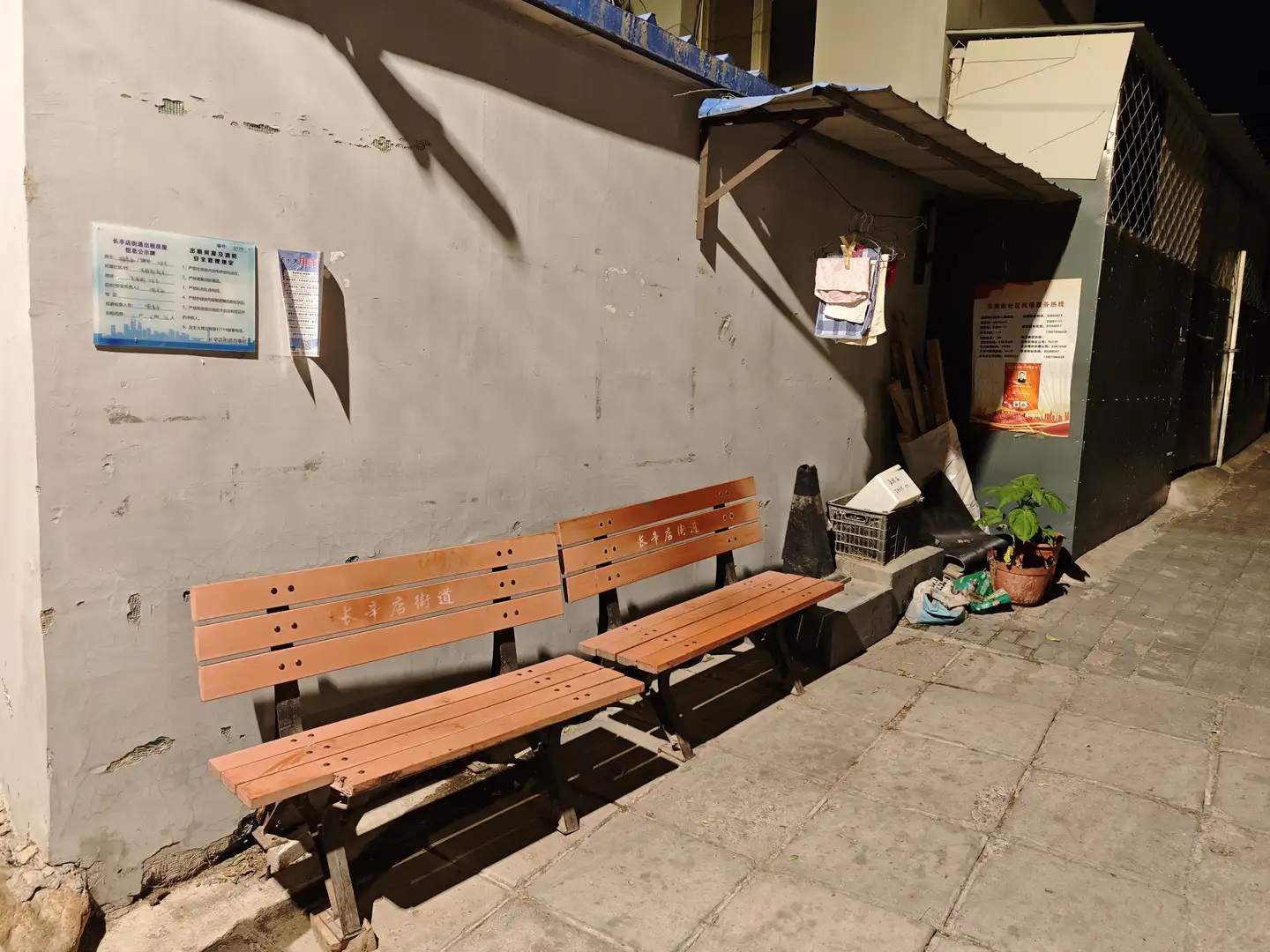 夜晚的街角,两排木质长椅靠墙放置,墙上贴有蓝色告示牌。长椅旁边有一个黑色交通锥和一些杂物。墙角处摆放着几盆绿色植物。