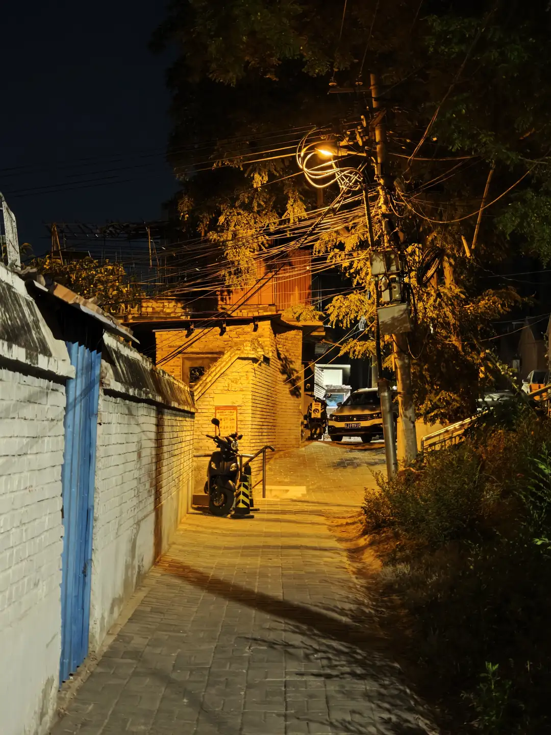 夜晚的小巷,路灯温暖的光芒洒在地面上,左侧是白色围墙和蓝色大门。右侧有树木和灌木丛,小巷尽头停放着一辆摩托车和汽车。电线在空中纵横交错。