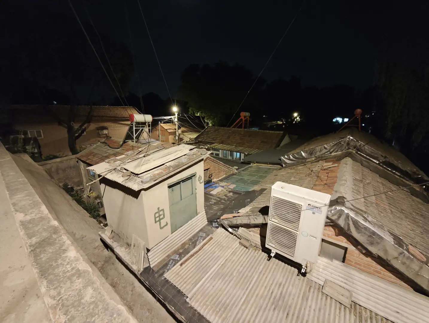 夜晚的屋顶视角,瓦片和金属板构成屋顶表面,上面放置着空调外机和储水罐。远处可以看到更多房屋和树木的轮廓。天空漆黑,几盏灯光点缀其间。