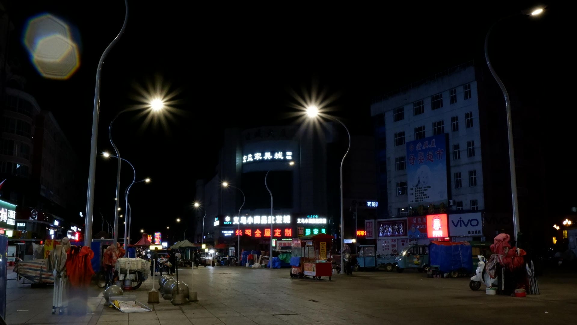 夜市广场上，路灯亮着，周围有摊位和广告牌。